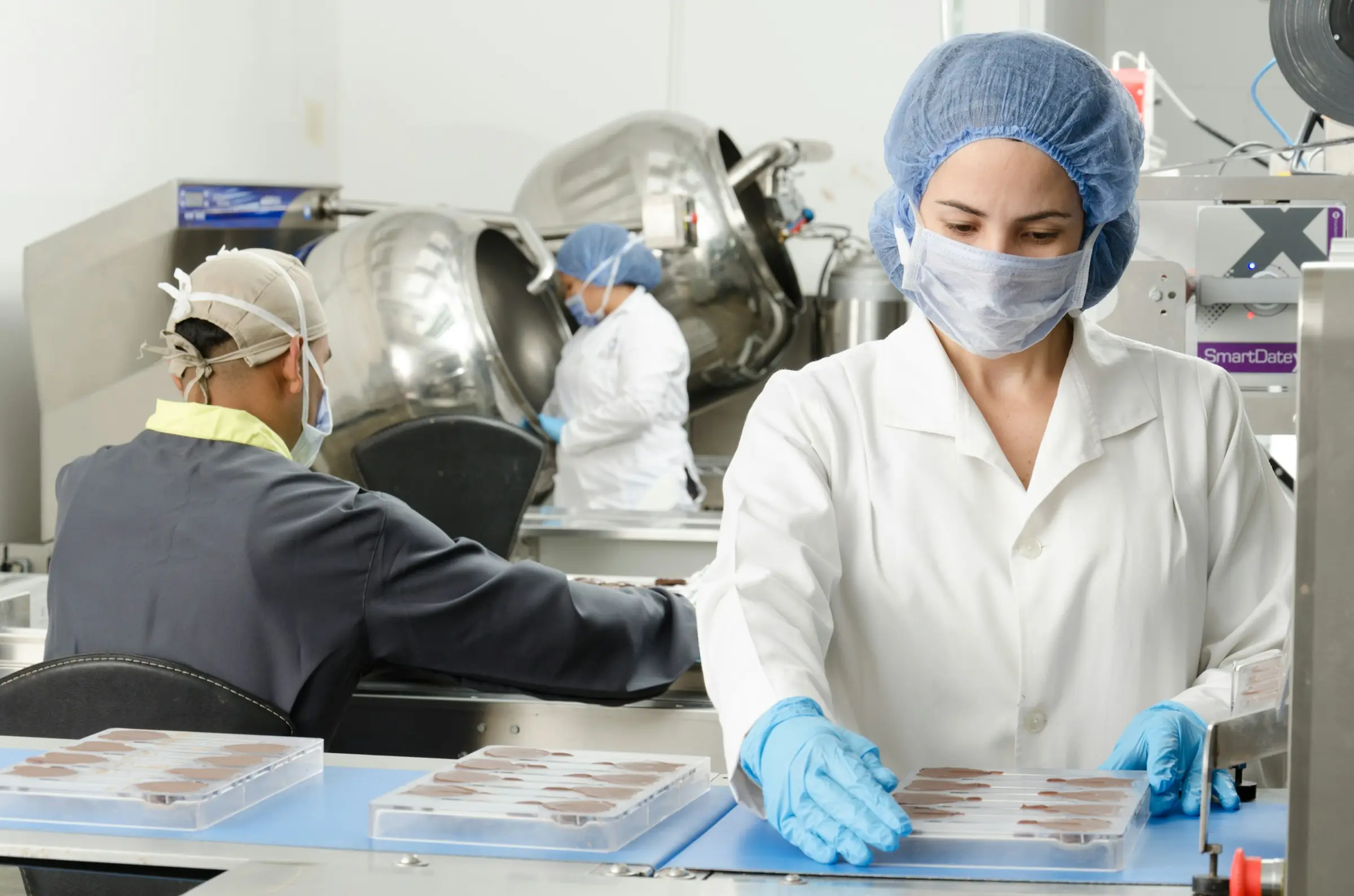 Eine Mitarbeiterin mit Haarnetz, Mundschutz und Handschuhen arbeitet an einer Produktionslinie.